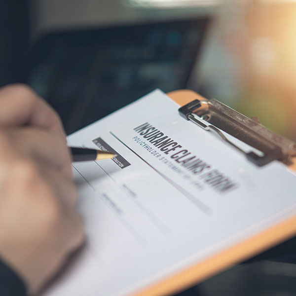 Hand holding pen writing insurance claims form clipboard office desk. Person filling policyholder statement paperwork medical auto health accident property coverage financial protection