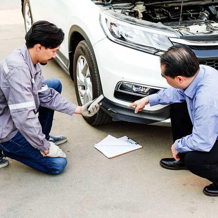 Mechanic and client kneel by a white car with the hood open, inspecting front bumper damage and discussing repairs beside a clipboard